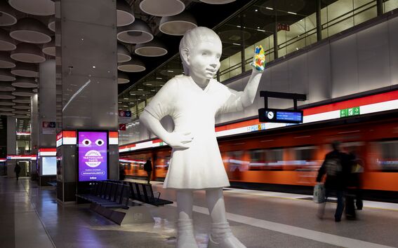 A view from Tapiola metro station, where there is a large white sculpture of a girl. She has raised her hand in greeting, her palm covered in different colors. An orange metro train passes by.