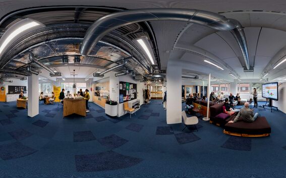 A modern office space with people working and discussing. Exposed ducts on the ceiling, blue carpet, and white walls.