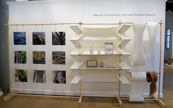 Näyttelyseinä, jossa luontovalokuvia, valkoiset origamihyllyt ja roikkuvia valkoisia sekä ruskeita kartonkirullia.