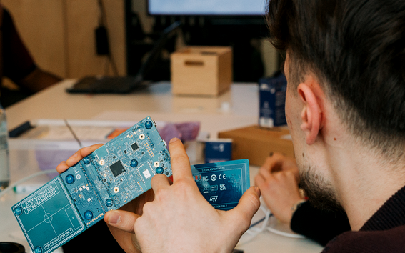 Two people at a desk, one holding blue circuit boards while discussing electronics