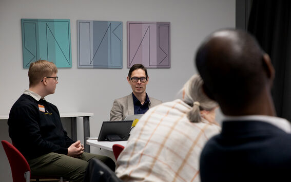 People in a meeting room talk, seated on red chairs, with pastel abstract art on the wall behind them.