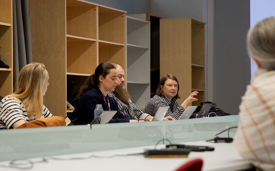 People in a meeting room sit at laptops, one speaking, with shelves and a projection screen behind.