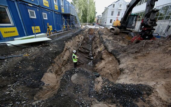 Worker in high-vis gear stands in deep trench at city construction site with blue cabins and excavator nearby