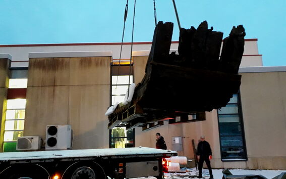 Cranes lift a large dark wooden wreck onto a snowy lorry beside an industrial building at dusk