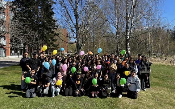 Large group of students outdoors holding colourful balloons on a sunny day, with leafless birch trees behind