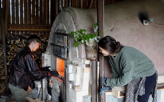Two people tend a glowing wood-fired kiln in a rustic wooden shed, with logs stacked beside them.