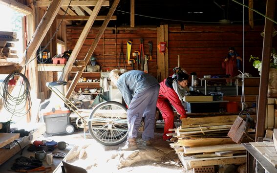 People working with wood and tools in a bright rustic workshop, shelves and planks around them