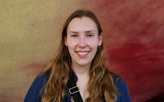 Smiling person with long hair and blue shirt, standing before a yellow and red textured wall