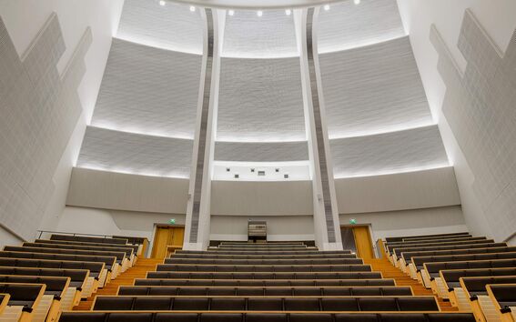 Bright modern lecture hall with grey tiered seats, pale wood steps and high curved white ceiling lights
