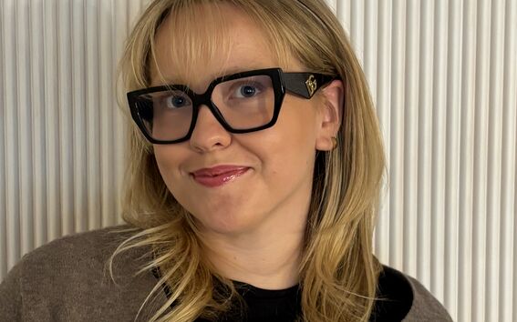 Smiling person with blonde hair and large black glasses, wearing a brown top against a light ribbed wall