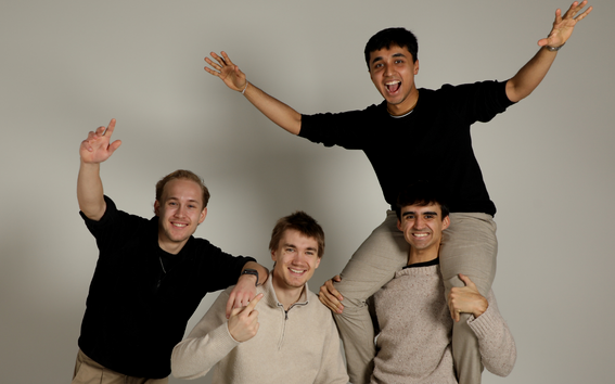 Four cheerful young men posing in a studio, one sitting on another’s shoulders, all smiling and waving