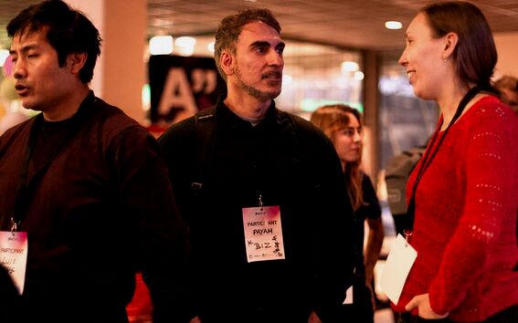 Three people with name badges talk at an indoor event, one in a red jumper, two in dark tops, with others behind.