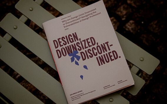 Esitteessä teksti ”Design, Downsized, Discontinued” vihreällä metallisella tuolilla, taustalla mukulakivet.