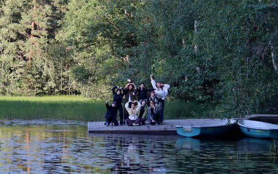 Group posing playfully on a wooden lakeside pier, green forest behind and two rowboats by the dock