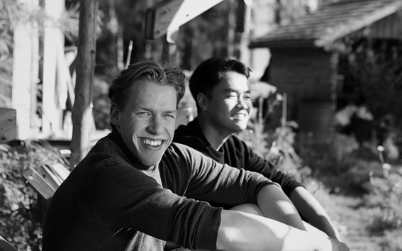 Two young men sit on a wooden bench outdoors, smiling in bright sunlight in a black and white photo