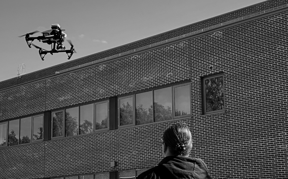Person with tied-back hair flying a dark drone by a large brick building with long windows