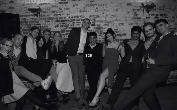 Group of friends in suits and dresses posing playfully with raised legs in front of a brick wall