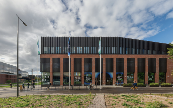 Modern brick and glass building with tall dark panels, street in front and people walking and cycling