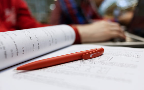 An Aalto pen lying on the page of a study book, students working in the background / photo by Aalto University, Aino Huovio