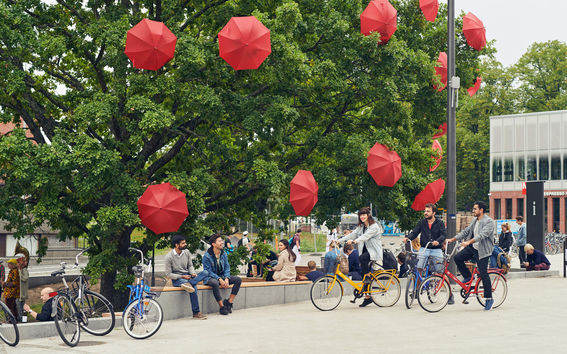 Students on Otaniemi campus. Open red umbrellas attached to trees, students sitting on a bench under the green-leaved trees. Photo by Aalto University /Unto Rautio