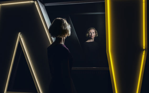 Kuvassa selin seisovan naisen hymyilevät kasvot näkyvät heijastumana peilistä 