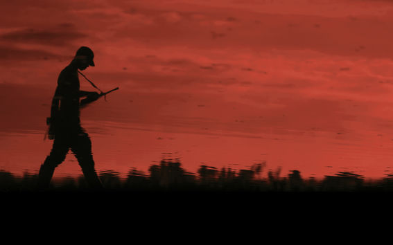 Tumma aseistautunut mieshahmo kävelee punaisen auringonlaskun maisemassa 