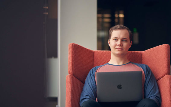 A male student sits in a red armchair and gazes into the distance with a Mac laptop on his lap