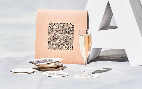 Kuvassa neliön muotoinen Äpy-lehti, kuohuviinilasi ja lasinalusia.