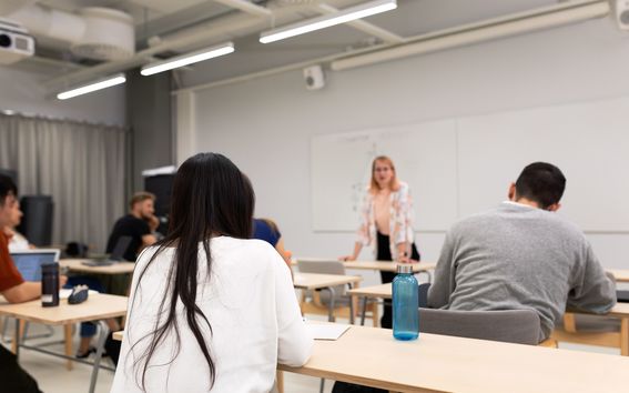 Opetus on käynnissä luokkahuoneessa ja opiskelijat kuuntelevat pöytään nojaavaa opettajaansa.