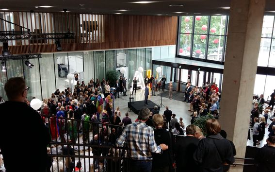 Joukko kerääntynyt moderniin rakennukseen isojen ikkunoiden ja puisten yksityiskohtien kanssa, seuraa puhujaa lavalla.