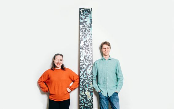 Tohtorikoulutettavat Noora Yau ja Konrad Klockars seisovat vierekkäin valkoista taustaa vasten. Heidän välissään on pystysuora puinen levy, joka on pohjaväriltään musta, mutta pinnassa on kirkkaita sinisiä ja keltaisia kuvioita. Kuva: Jaakko Kahilaniemi.