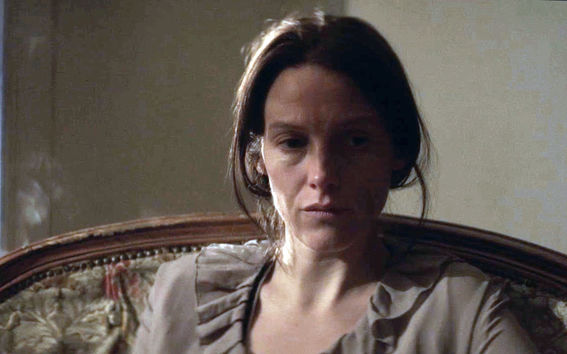 Mietteliään ja hieman surullisen näköinen nainen istuu lähikuvassa sohvalla ja katsoo alaviistoon, taustalla vaalea seinä.