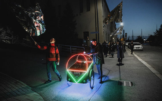 A photo of a performance where a person is pushing a two-wheeled cart in the street. The cart is lit with neon lights, around are people who are waving silver flags.