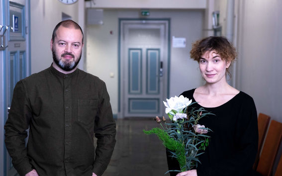 Mies seisoo kädet taskuissa, naisella on kukkia kädessään, molemmat tummissa vaatteissa seisovat käytävällä.