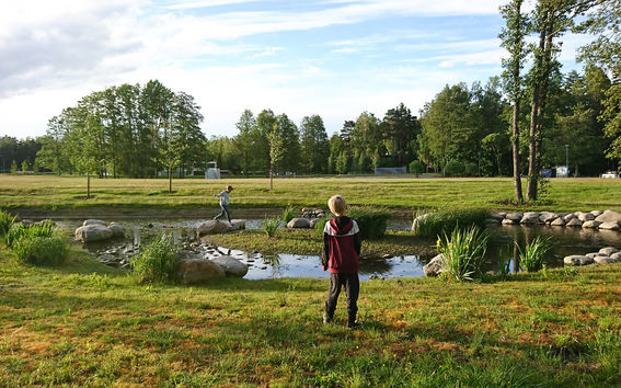 Kaksi poikaa leikkimässä ilta-auringossa kaupungin viheralueella, jonka keskellä on pieni lampi ja sen ympärillä kiviasetelma.