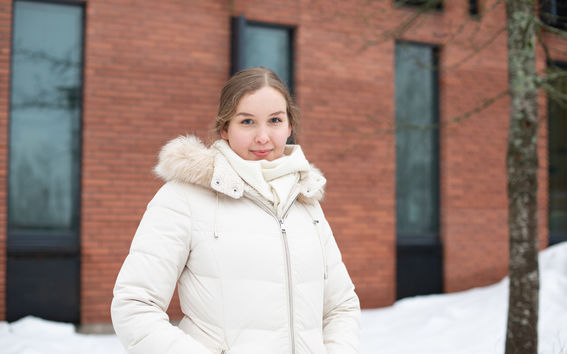 Heli Julkunen seisoo ulkona ja katsoo hymyillen kameraan. Taustalla näkyy oranssinruskea tiilirakennus ja lumihankia.