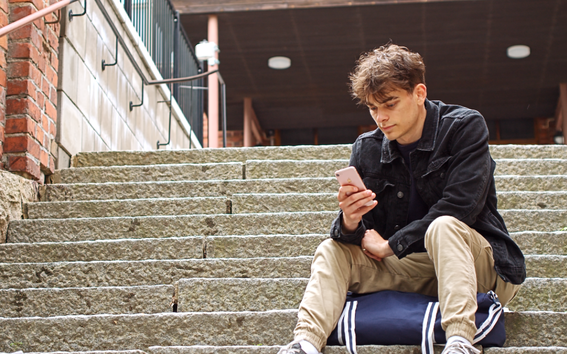 Nuori mies istuu rakennuksen kiviportailla ja selailee puhelintaan.