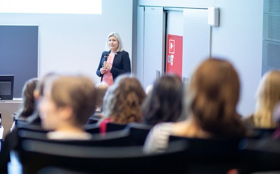 Nainen puhuu täydelle luentosalille, yleisöstä näkyy takaraivot