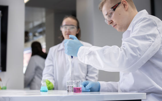 Two people working in a laboratory. Both are wearing lab coats as well as protective glasses and gloves.