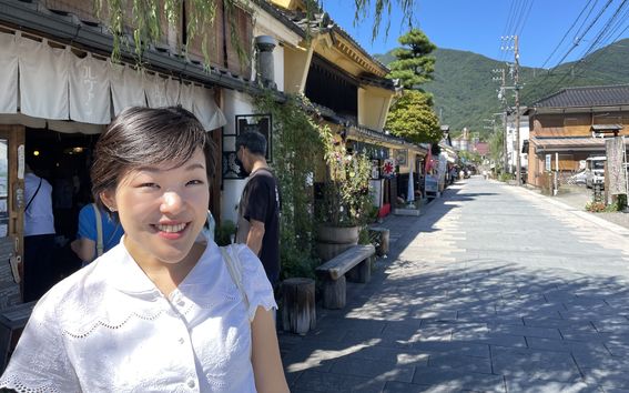 Chisako Ogiwara Digital Business Master Class alumna from Aalto University Summer School standing outside a restaurant at a street with some lush and green mountains in the background. She is wearing a short-sleeved white blouse and is smiling towards the camera.