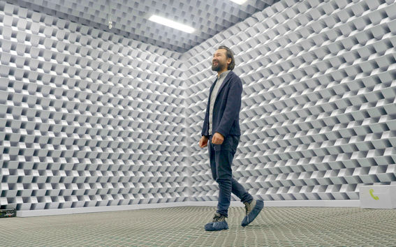 A man in a dark suit stands in an anechoic chamber with grey, pyramid-shaped foam panels.