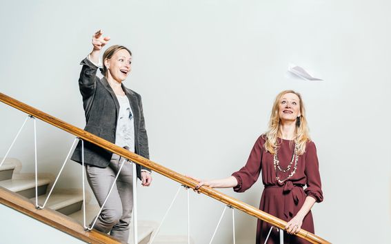 Marjo-Riitta Diehl ja Miisa Mink seisovat vaaleassa portaikossa rappusilla. Diehl on heittänyt ilmaan paperilennokin, joka leijailee Minkin kasvojen yläpuolella. Molemmat hymyilevät. 