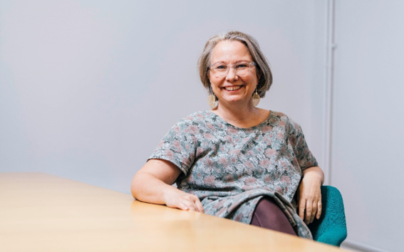 Laura Arpiainen in a patterned dress, sitting on a green chair and leaning against a table, smiling happily towards the camera.