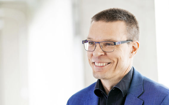 Keski-ikäinen mies hymyilee. Hänellä on sininen pikkutakki ja mustasankaiset silmälasit.