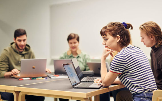 Opiskelijoita opiskelemassa tietokoneilla pöydän ääressä.