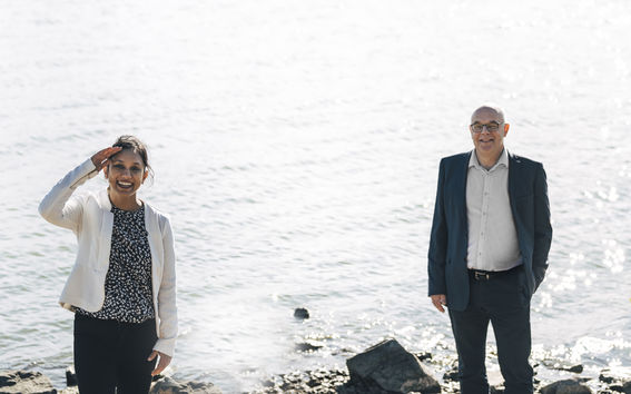 Mashrura Musharraf ja Sauli Eloranta seisovat aurinkoisena päivänä merenrannassa. Etualalla näkyy rantakiviä, taustalla meri. Musharraf tervehtii kuvaajaa hymyillen kuten laivan kapteeni, "vieden kättä lippaan".