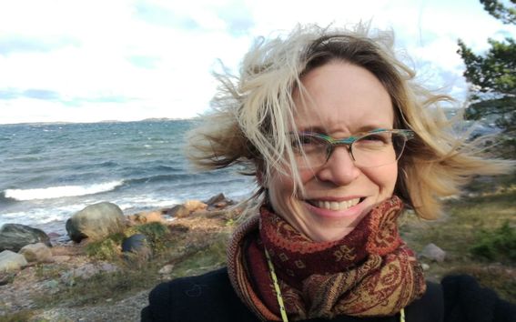 Nainen rannalla tuulisella säällä hymyilee, taustalla meri kuohuaa 