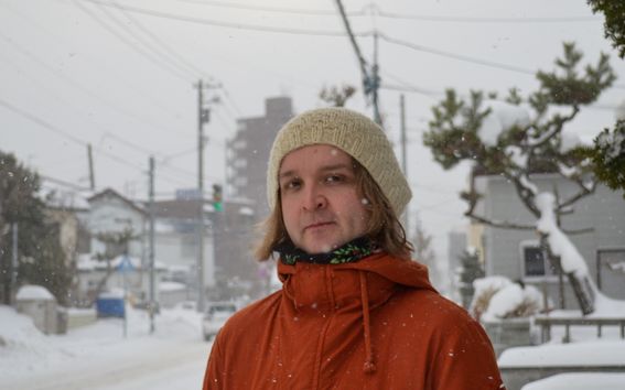 Mies punaisessa takissa pipo päässä seisoo kädet taskuissa lumisessa maisemassa