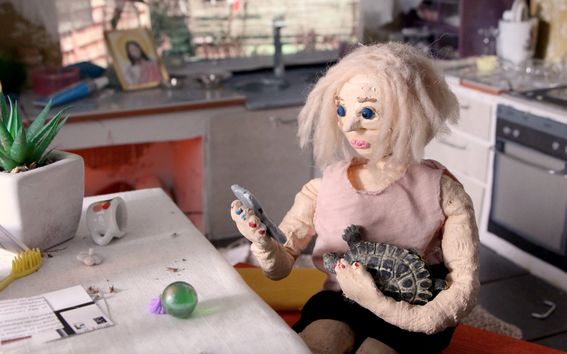 Äiti-nukkehahmo istuu pöydän ääressä lavastetussa ympäristössä matkapuhelin kädessään