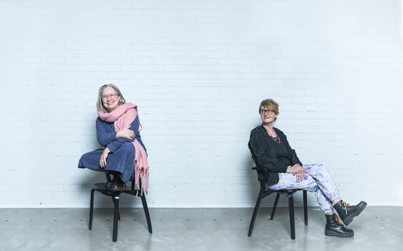 Laura Arpiainen ja Susanna Helke istumassa mustilla tuoleilla valkoisen tiiliseinän edessä. Arpiainen istuu tuolin selkänojan päällä jalat istuinosalla ja Helke katsoo häntä hymyillen.